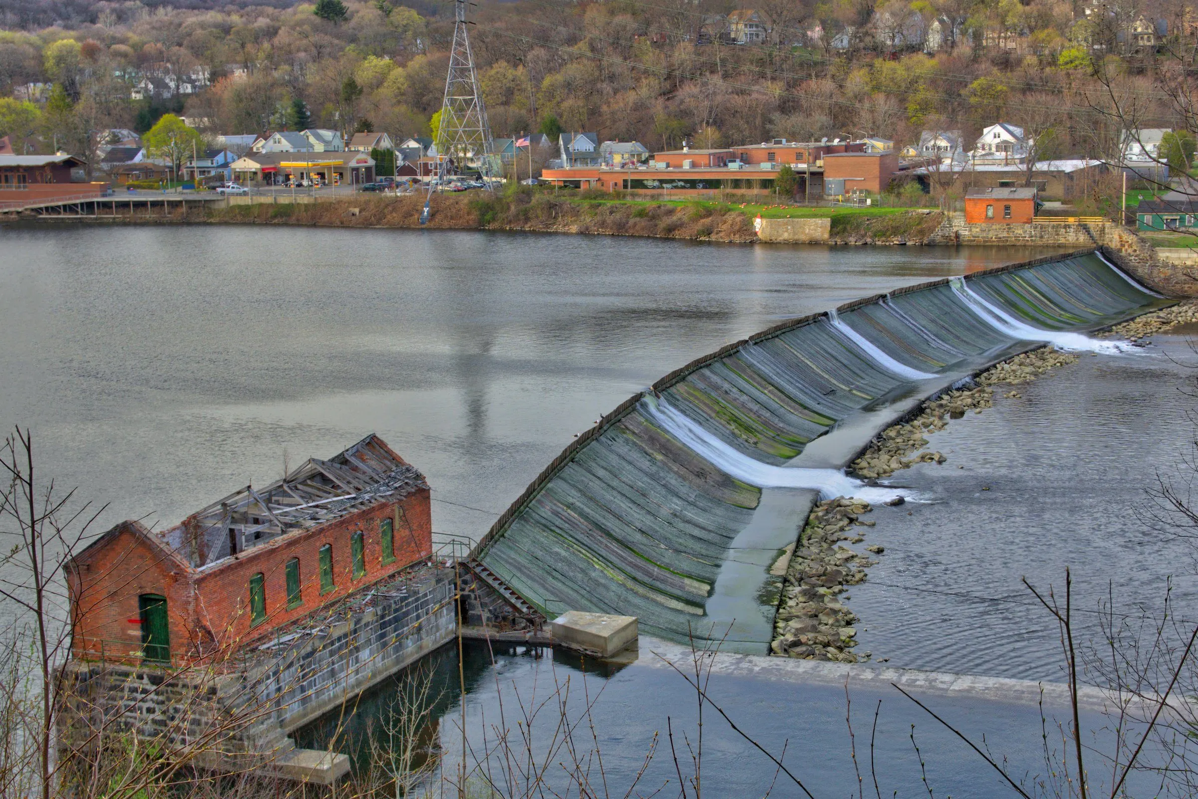 The curved Ousatonic Dam on the Housatonic River, with the old brick powerhouse in front and the Shelton riverfront and hillside across the water
