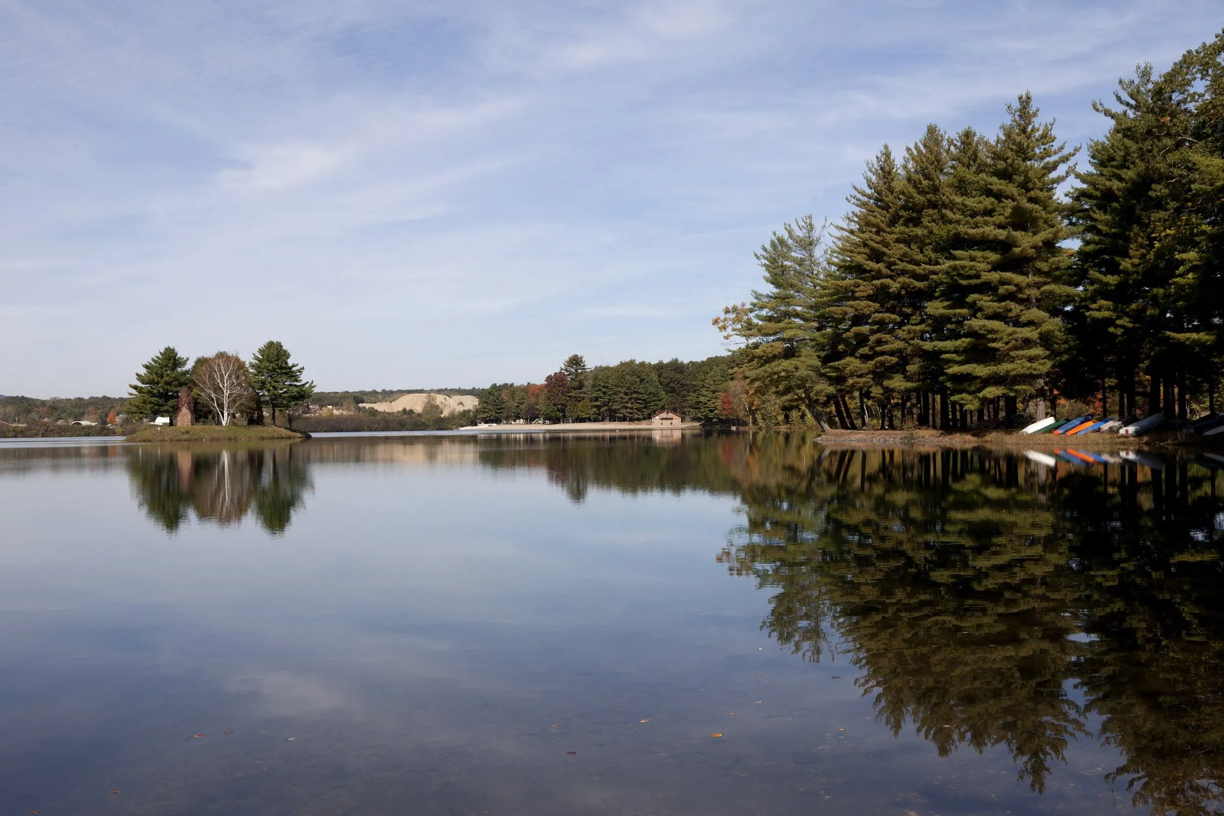The beach and boathouse of Dunning Lake under tall pines, reflected in still water