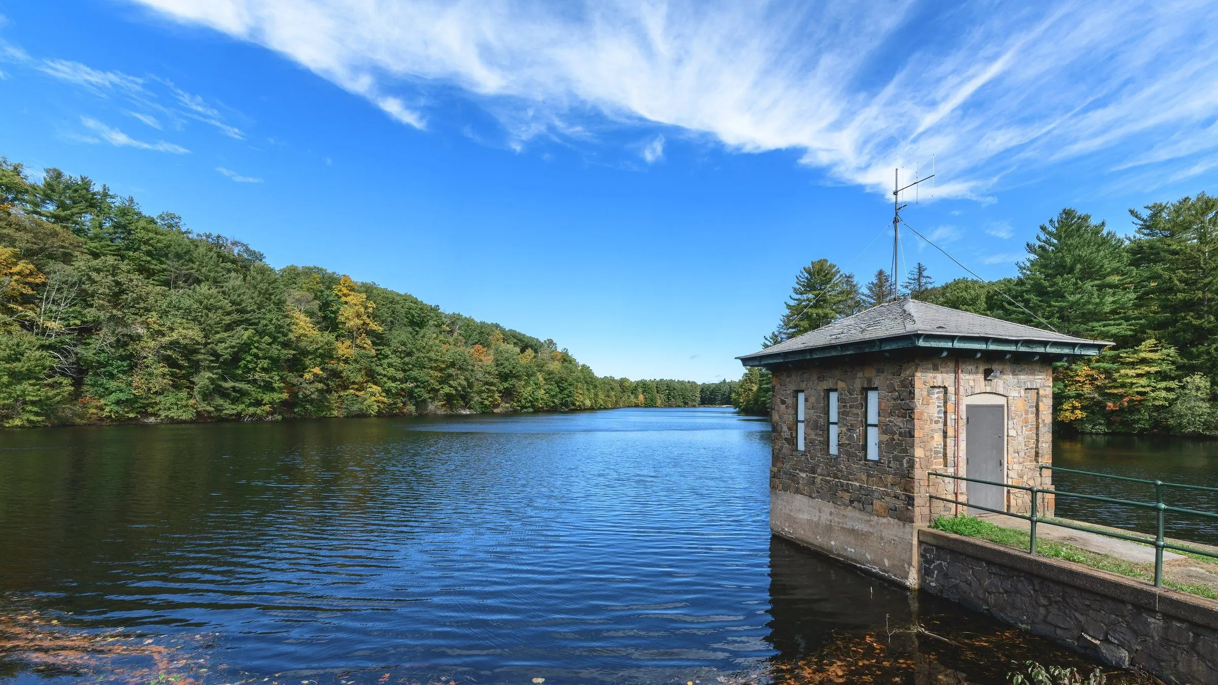 A stone gatehouse on the West Hartford Reservoir, with wooded hills and a bright blue sky