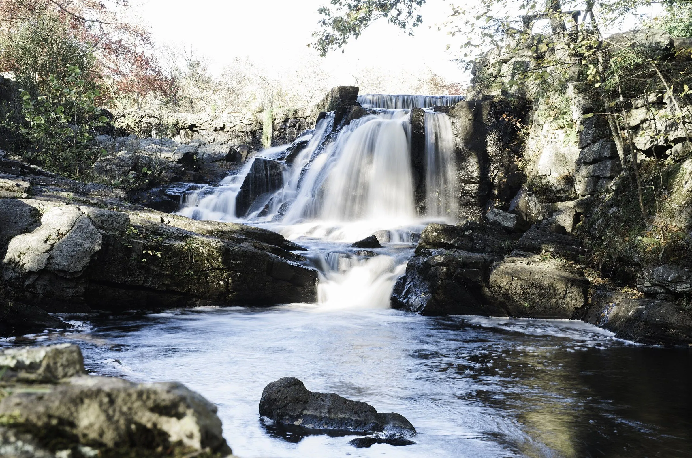 Southford Falls stepping down over dark rock ledges into a pool, with autumn trees and old stone walls