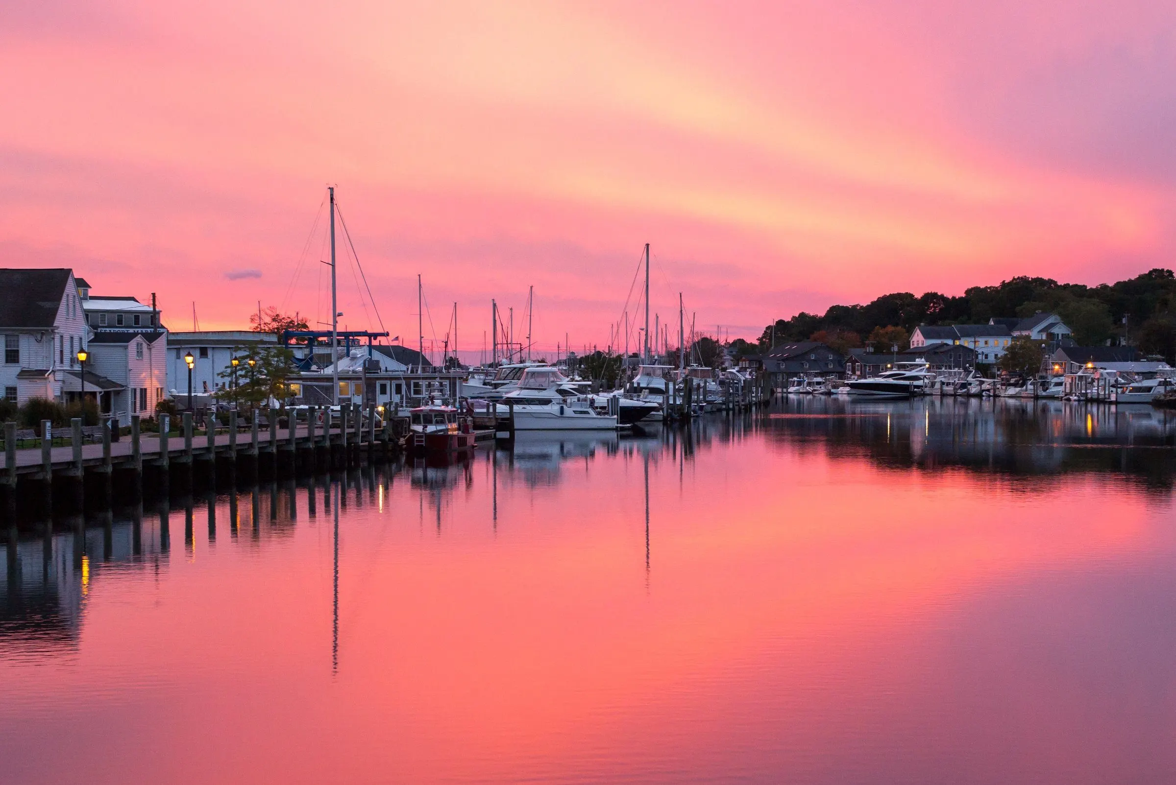 Boats and waterfront buildings along the Mystic River, reflected in water lit pink by the sunrise